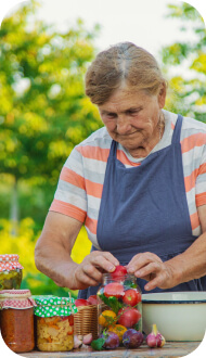 пожилая женщина в футболке с серыми, оранжевыми и белыми полосками, в синем фартуке, складывает красные помидорки в банку, рядом стоят ещё несколько банок уже закрытые цветной бумагой и перевязаны верёвочкой